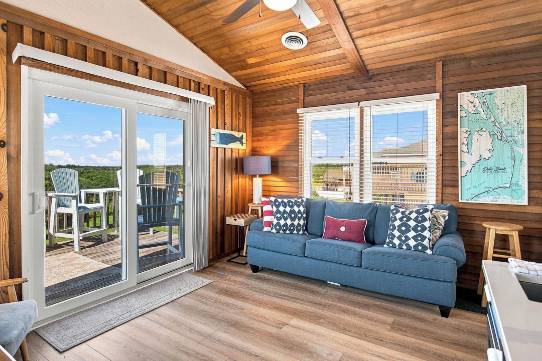 A cozy living room with a blue sofa, a wooden ceiling, and a large sliding glass door leading to an outdoor deck.