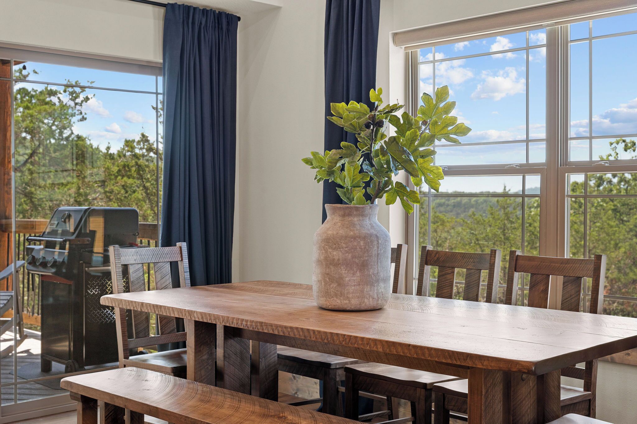 A wooden dining table with chairs, a vase with green foliage, and a grill outside.