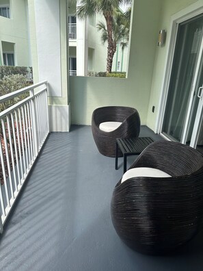 A balcony with two wicker chairs and a small table, a white railing, and a view of a building with balconies and a palm tree. - Modern Luxury one bedroom apartment in Doral Florida! (Doral)