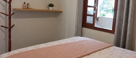 A bedroom with a bed, a wooden coat rack, a shelf with toiletries, and a window with curtains.