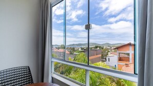 A balcony with a view of a residential area, a metal railing, and a window with a view of houses and hills. - Complete Studio 900 m From Lagoa Ecm0206 (Florianópolis)