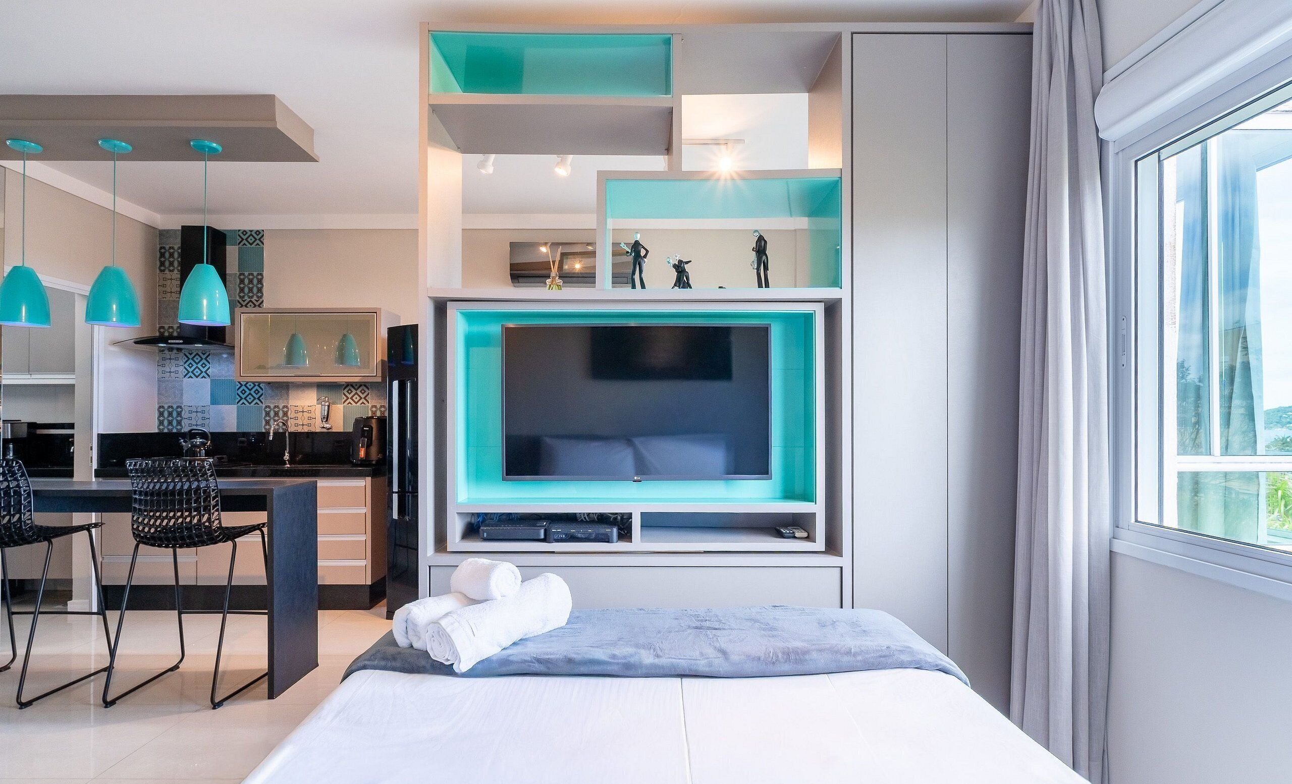 A modern hotel room with a large bed, a TV, a desk with chairs, and a kitchen area.
