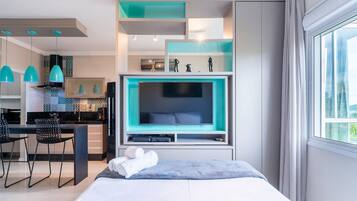 A modern hotel room with a large bed, a TV, a desk with chairs, and a kitchen area.