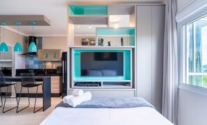 Una habitación de hotel moderna con una cama grande, televisión, escritorio con sillas y zona de cocina.