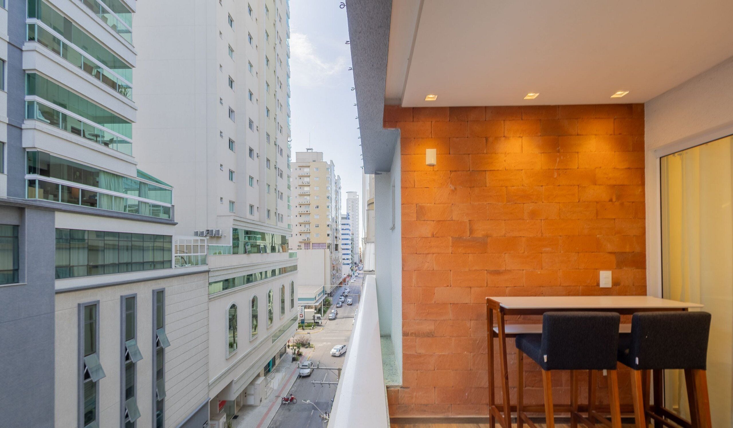 Uma varanda com mesa e cadeiras, com vista para uma rua da cidade com prédios e carros estacionados.