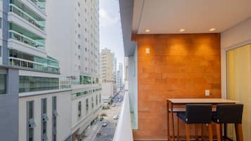 Uma varanda com mesa e cadeiras, com vista para uma rua da cidade com prédios e carros estacionados.