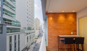 Uma varanda com mesa e cadeiras, com vista para uma rua da cidade com prédios e carros estacionados.