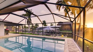 An indoor swimming pool with a glass enclosure, surrounded by palm trees and a covered seating area. - VILLA RIVERSIDE– HEATED POOL | GULF ACCESS | BOAT RENTAL + TOURS VOUCHER INCL. (Cape Coral)