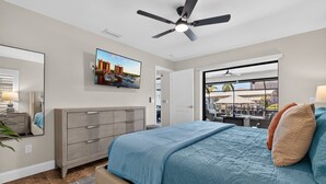 A bedroom with a bed, a dresser, a ceiling fan, a TV mounted on the wall, and a window with a view of an outdoor area. - VILLA RIVERSIDE– HEATED POOL | GULF ACCESS | BOAT RENTAL + TOURS VOUCHER INCL. (Cape Coral)