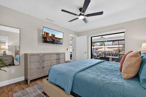 Ein Schlafzimmer mit Bett, Kommode, Deckenventilator, Wand-Fernseher und einem Fenster mit Blick auf einen Außenbereich.
