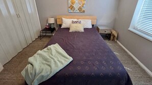 Een slaapkamer met een bed, nachtkastje, lamp, raam met zonwering en een deur.