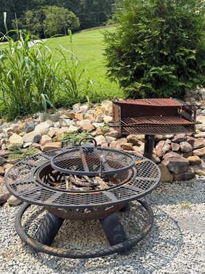 A metal fire pit with a circular design, surrounded by rocks and gravel, set in a garden with grass and trees in the background.