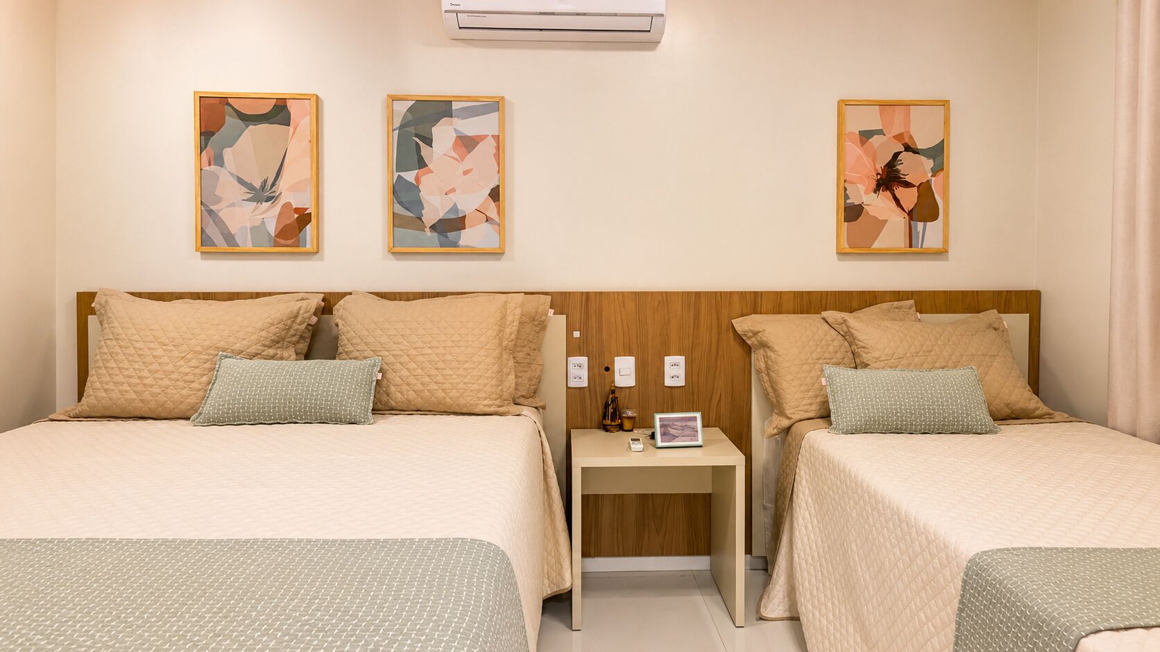 Quarto de hotel com duas camas, uma mesinha de cabeceira e quadros nas paredes.