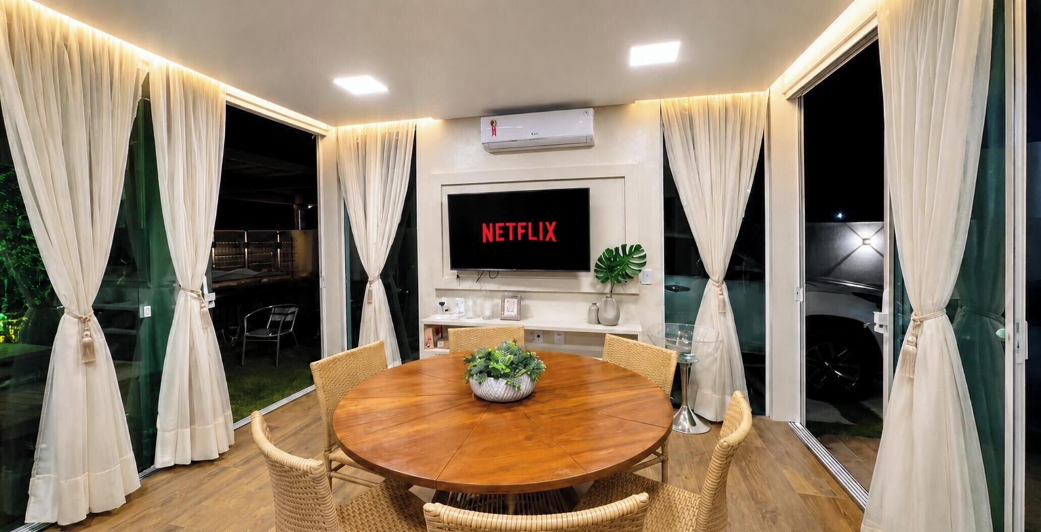 A dining area with a round wooden table, wicker chairs, a potted plant, and a wall-mounted TV displaying 'NETFLIX'.