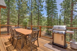 Eine Terrass mit Holztisch und -stühlen, einem Grill und Blick auf einen Wald.