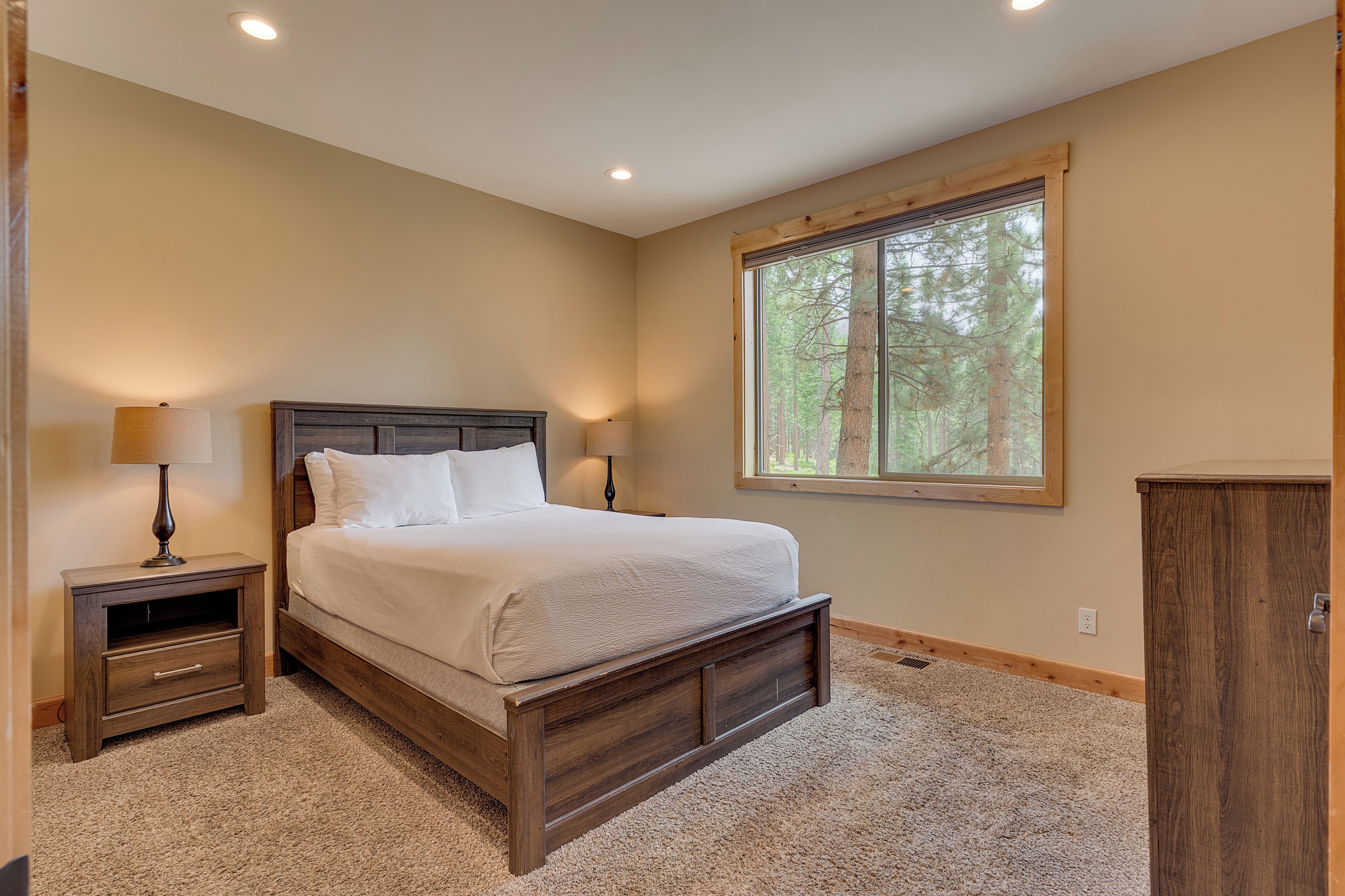 A bedroom with a wooden bed, two bedside tables with lamps, a large window with a view of trees, and a carpeted floor.