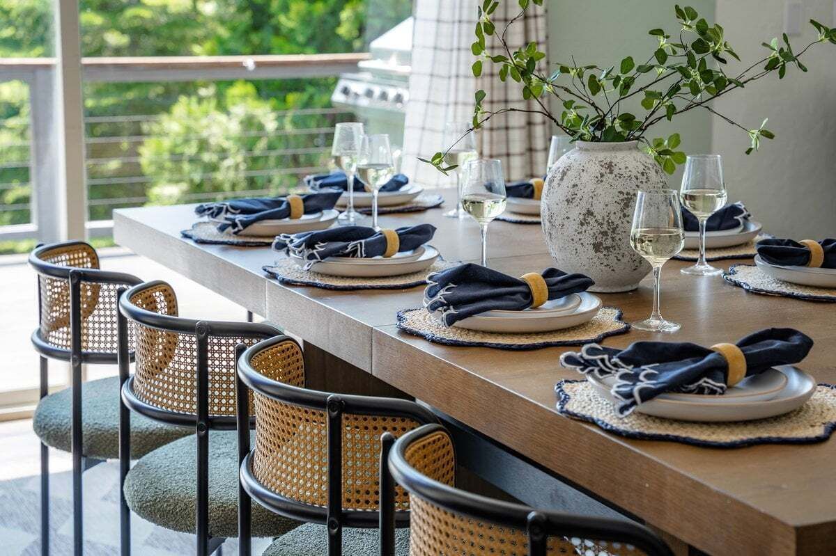 Una mesa de comedor dispuesta con platos, vasos y servilletas, rodeada de sillas de mimbre.