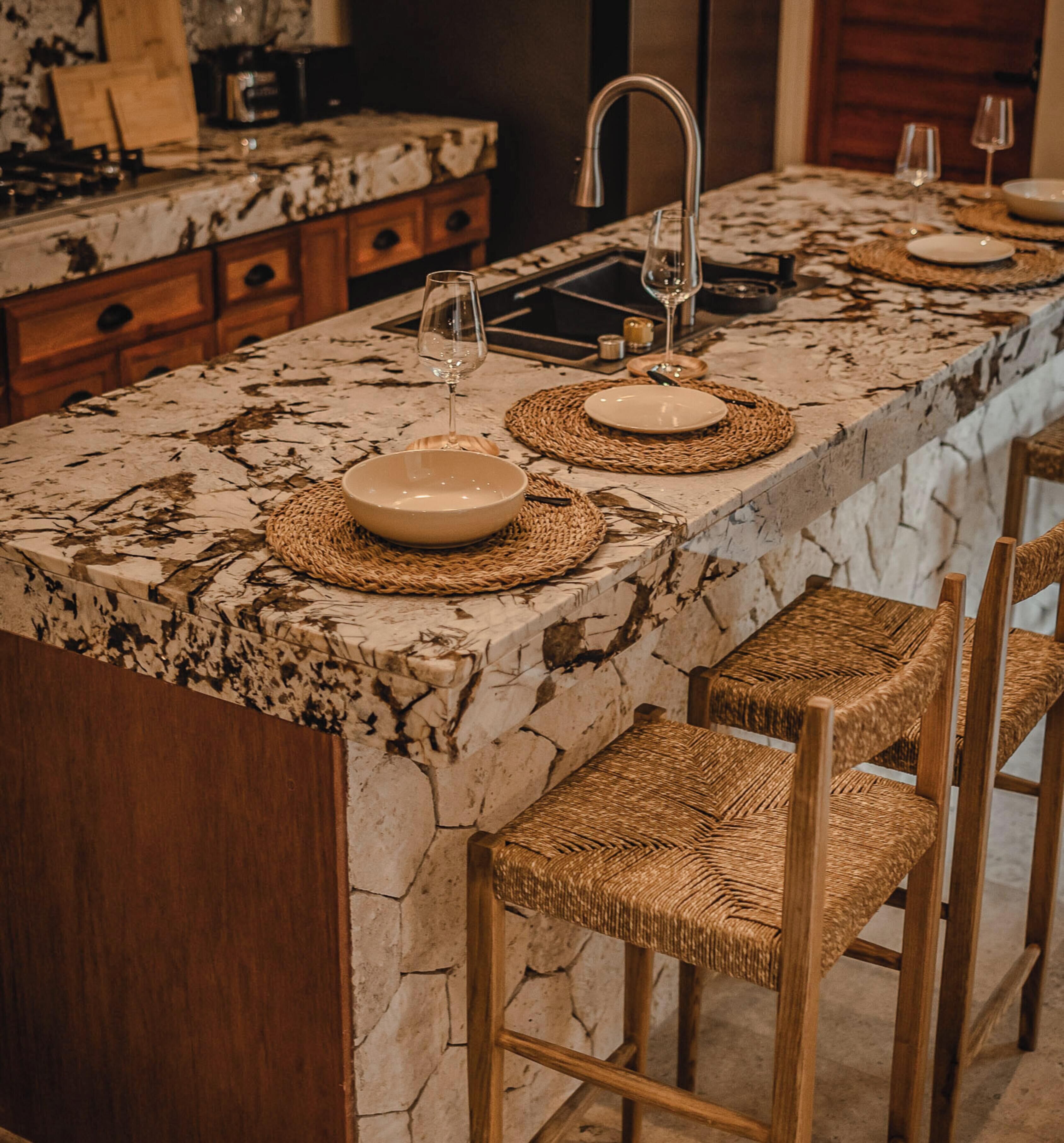 A kitchen island with a stone countertop, two bowls, a wine glass, and wicker chairs.