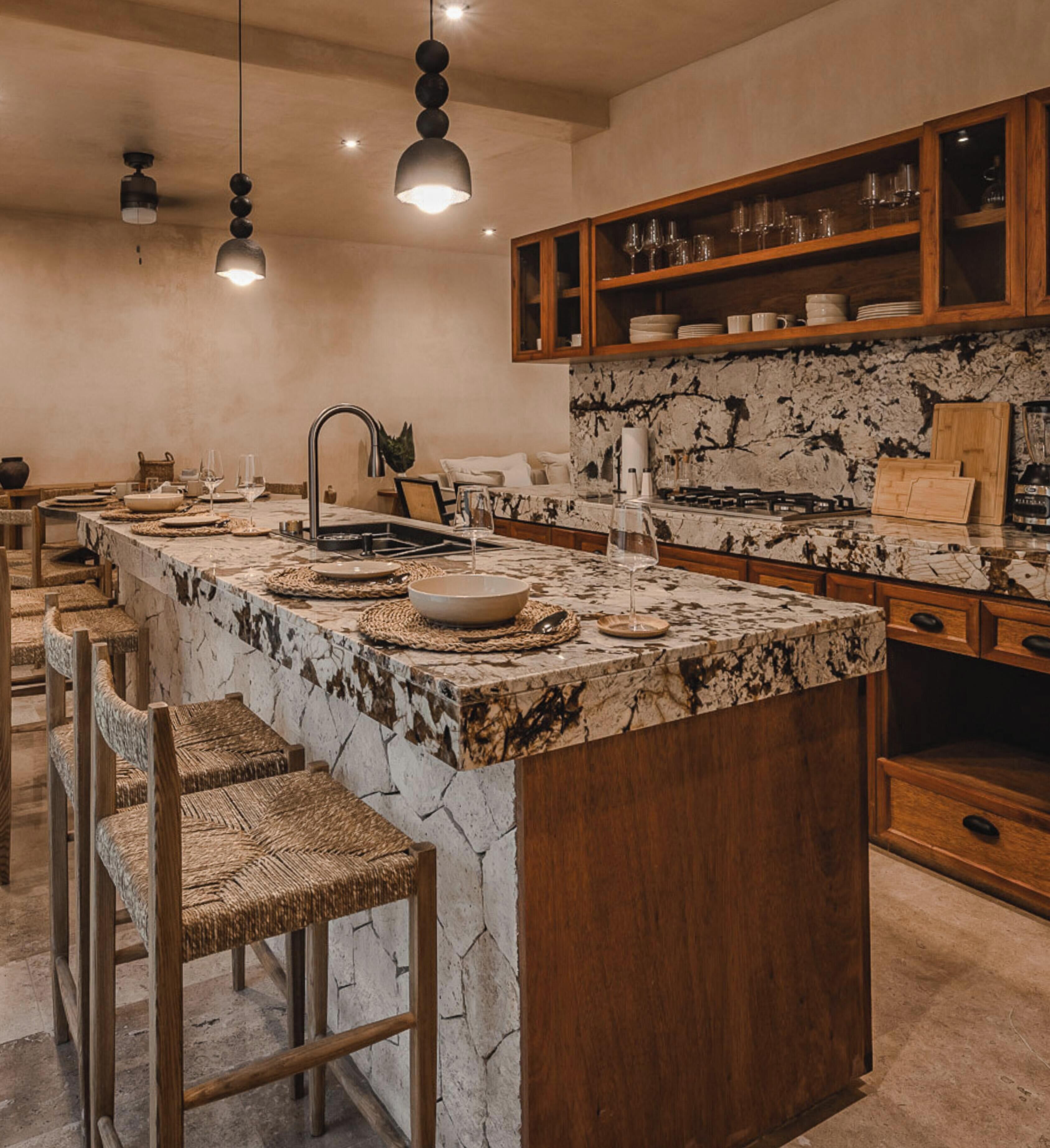 A kitchen with a marble island, wooden bar stools, and a wooden upper shelf with glassware.