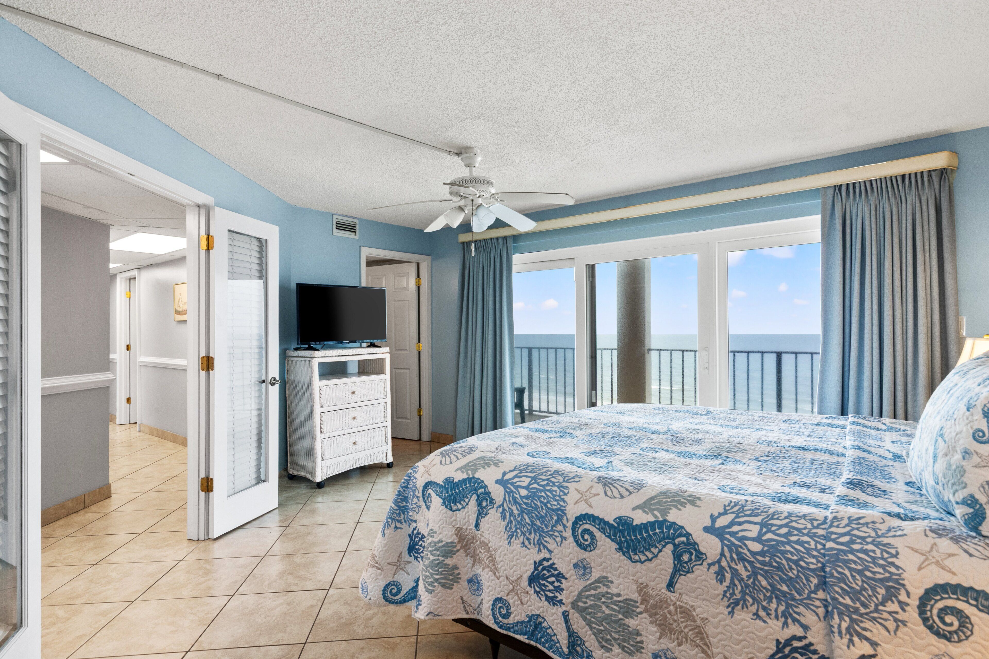 A hotel room with a bed, a television, a dresser, and a view of the ocean.