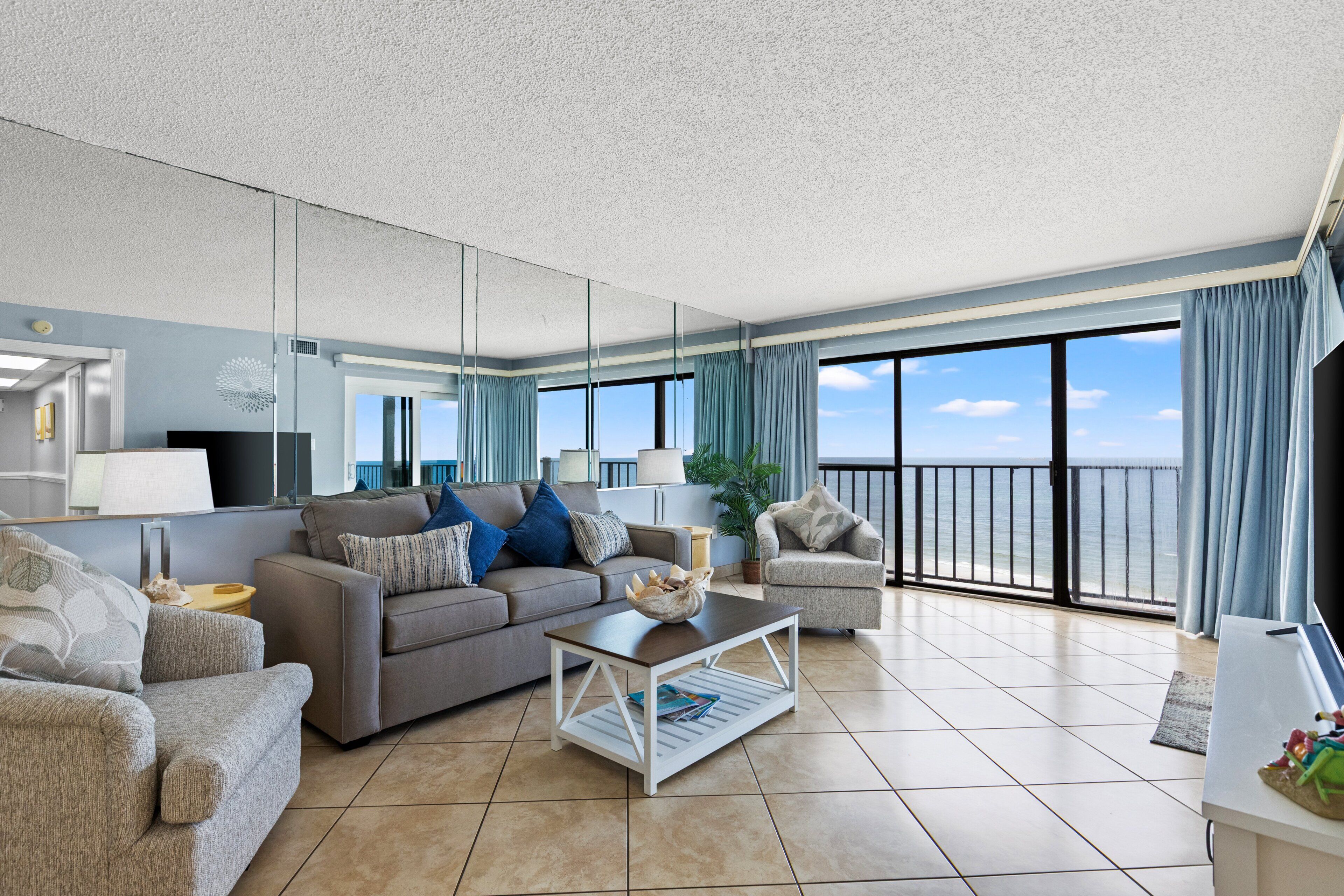 A modern living room with a sofa, armchair, and coffee table, a large window with a view of the sea, and a balcony with a railing.