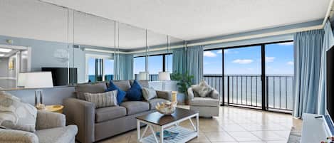 A modern living room with a sofa, armchair, and coffee table, a large window with a view of the sea, and a balcony with a railing.