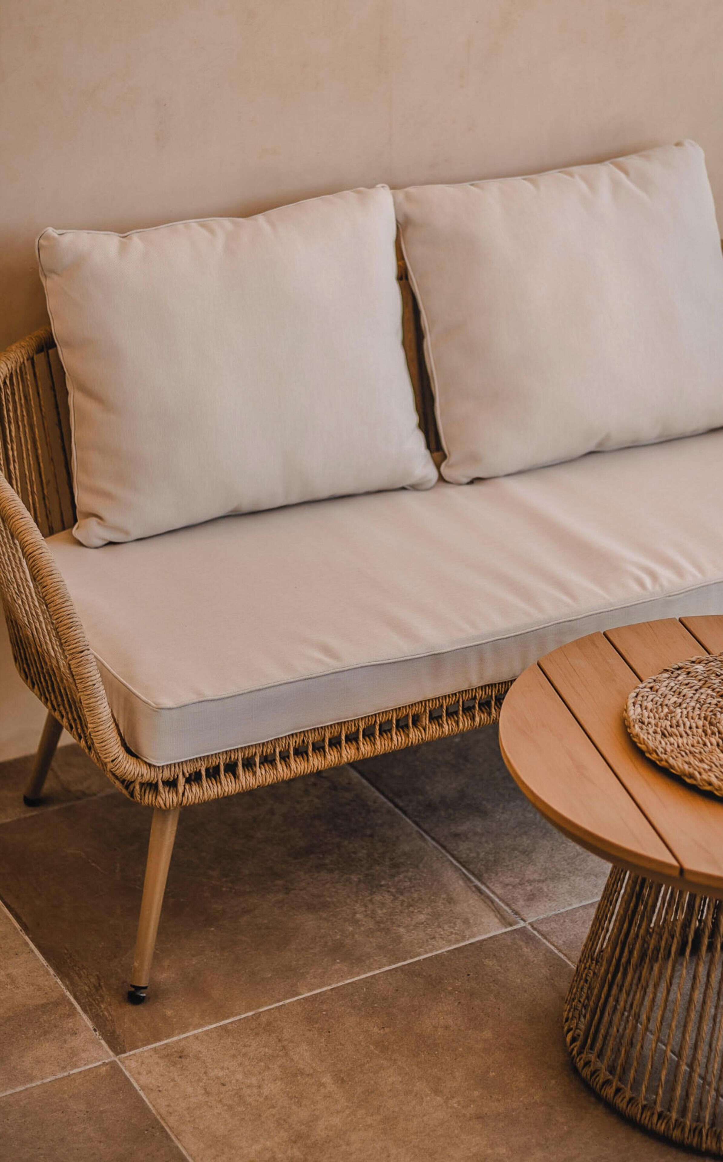 A wicker sofa with cushions against a plain wall, a woven table with a round woven placemat, and tiled flooring.