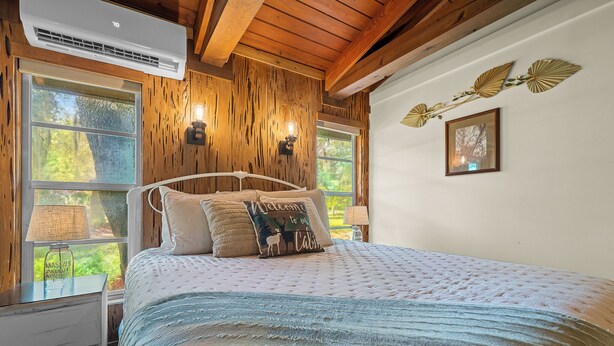 Ein Schlafzimmer mit Holzbalkendecke, einem Bett mit gemustertem Überwurf und einem Fenster mit Blick auf Grünflächen.
