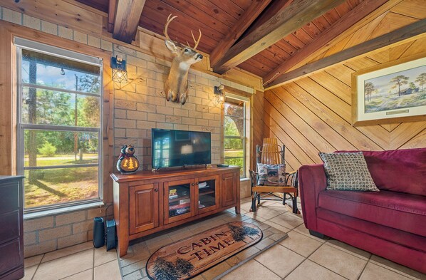 A cabin interior with a television on a wooden cabinet, a deer head mounted on the wall, and a red couch.
