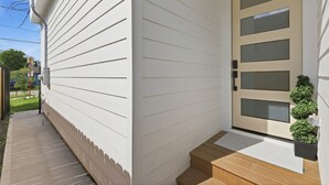 A house with a white exterior, a front door with glass panels, a small wooden staircase leading to the door, and a potted plant on the right side. - New Build Modern 3BR HTX Home • King Beds • Parking • Pet Friendly (Houston)
