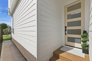 A house with a white exterior, a front door with glass panels, a small wooden staircase leading to the door, and a potted plant on the right side. - New Build Modern 3BR HTX Home • King Beds • Parking • Pet Friendly (Houston)