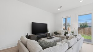 A modern living room with a large flat-screen TV, a sectional sofa, a coffee table, and a window with a view of a backyard. - New Build Modern 3BR HTX Home • King Beds • Parking • Pet Friendly (Houston)
