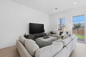 A modern living room with a large flat-screen TV, a sectional sofa, a coffee table, and a window with a view of a backyard. - New Build Modern 3BR HTX Home • King Beds • Parking • Pet Friendly (Houston)