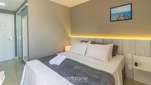Una habitación de hotel con una cama bien hecha, un cuadro de un muelle y una mesita de noche con una lámpara.