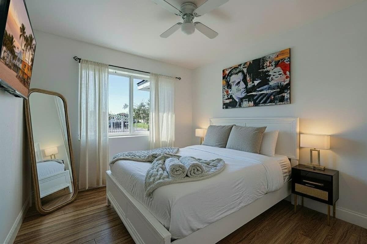 A bedroom with a bed, a nightstand, a ceiling fan, a window with curtains, and a painting on the wall.