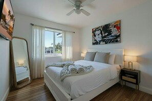Une chambre à coucher avec un lit, une table de chevet, un ventilateur de plafond, une fenêtre avec des rideaux et un tableau au mur.