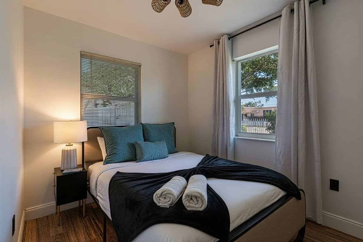 A bedroom with a bed, a nightstand, a window with curtains, and a view of the outside.