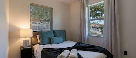 Un dormitorio con cama, mesita de noche, ventana con cortinas y vistas al exterior.