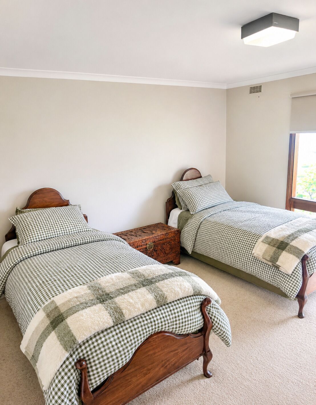 Un dormitorio con dos camas de madera, ropa de cama a cuadros, un pequeño cofre de madera y una lámpara en el techo.