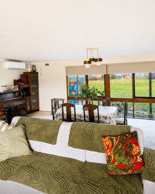 A living room with a sofa, dining table, chairs, and a view of a green field through large windows.