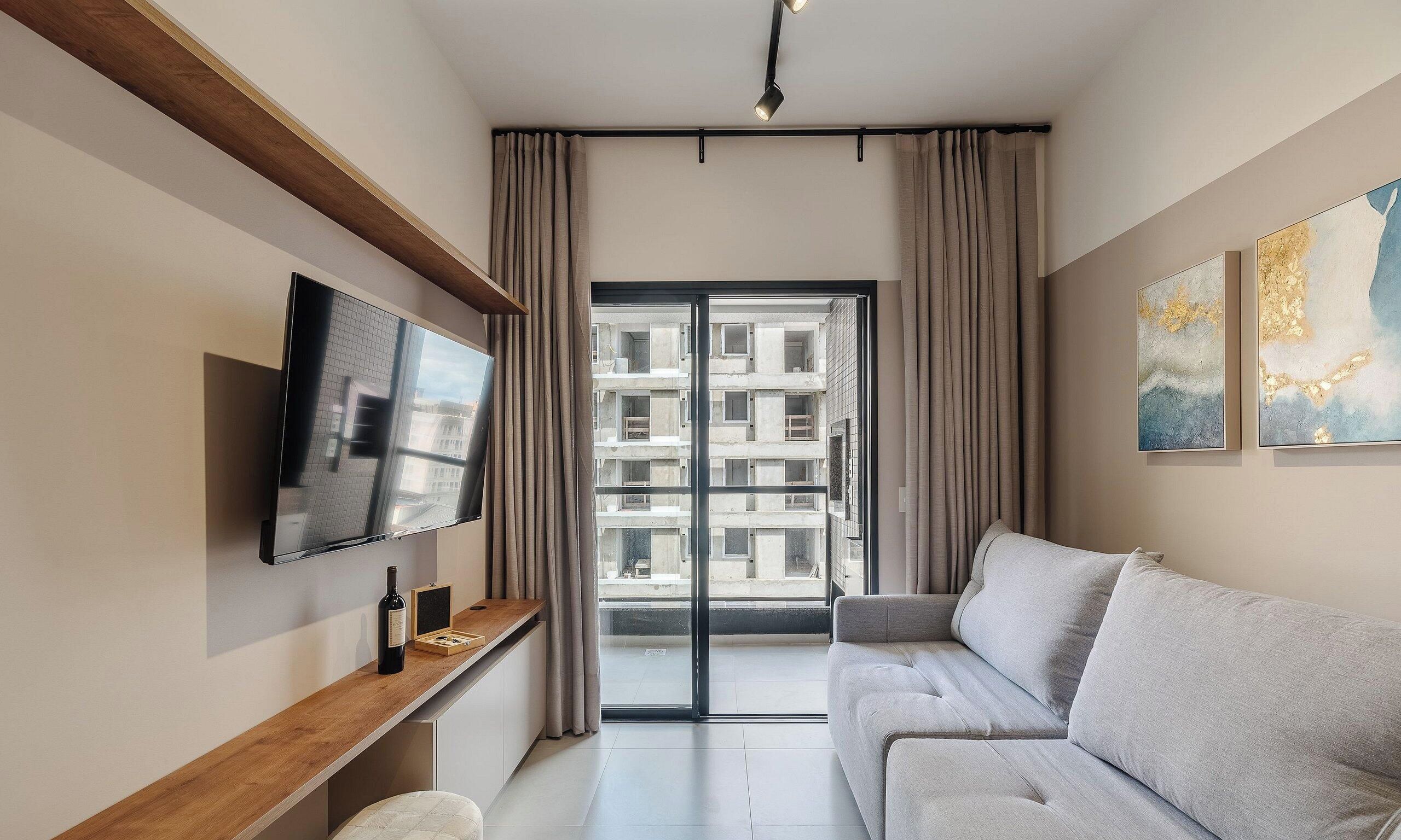 A modern living room with a flat-screen TV, a grey sofa, a wooden shelf, and a large window with curtains.