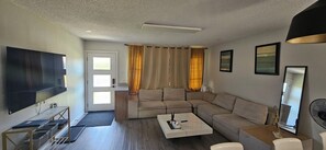 Ein Wohnzimmer mit einer Eckcouch, einem Couchtisch, einem Fernseher und einem großen Fenster mit Vorhängen.