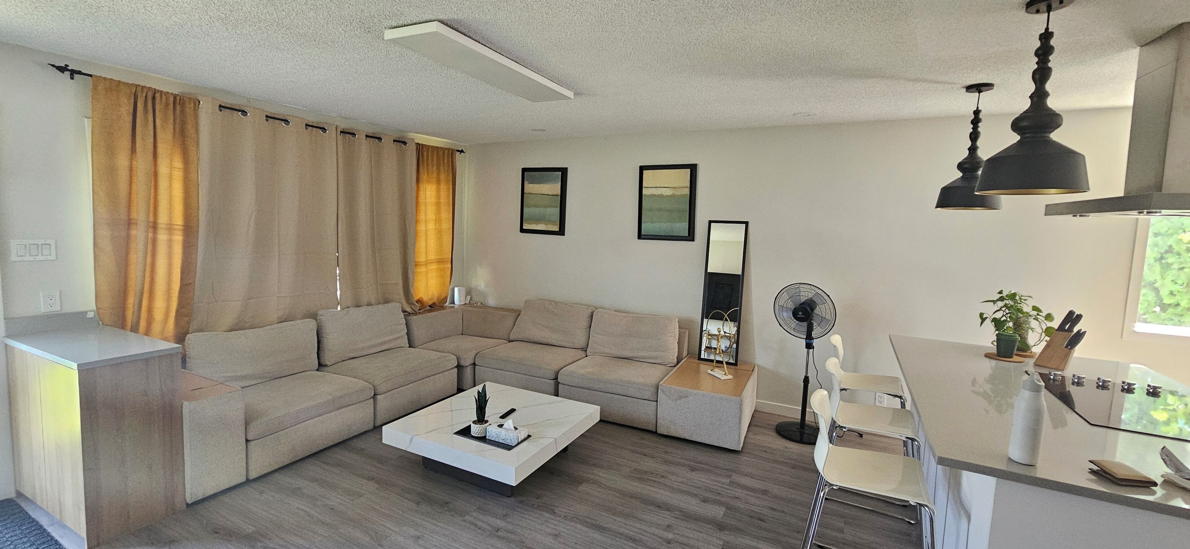 A modern living room with a sectional sofa, a coffee table, a standing fan, and a kitchen area with a countertop and a window.