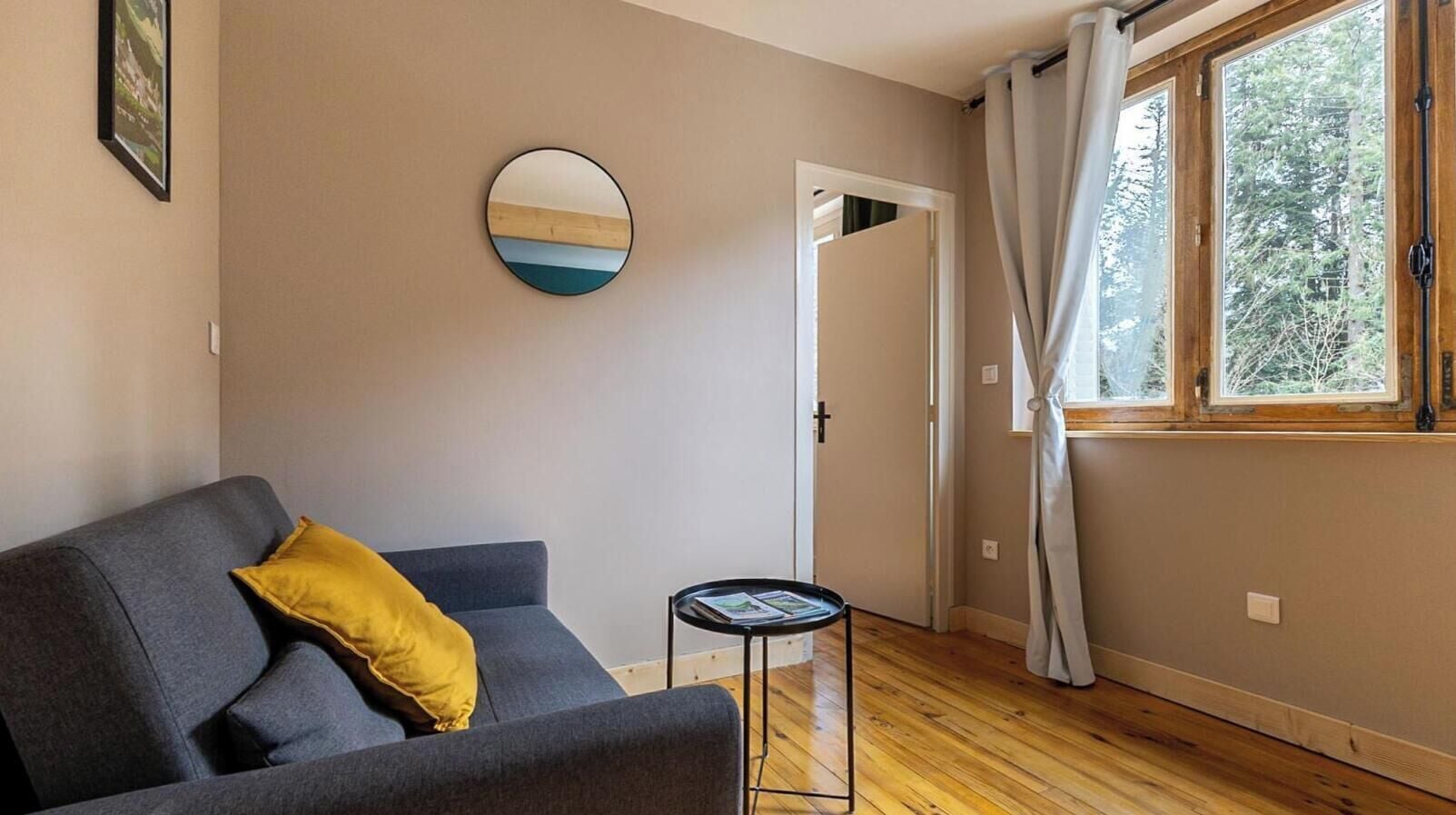 Une pièce avec un canapé gris, une petite table ronde, une porte et une fenêtre avec des rideaux.