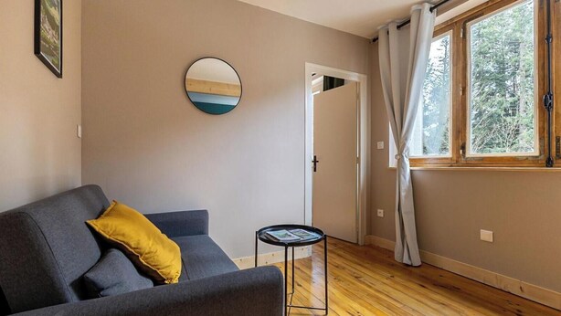Habitació amb un sofà gris, una taula rodona petita, una porta i una finestra amb cortines.