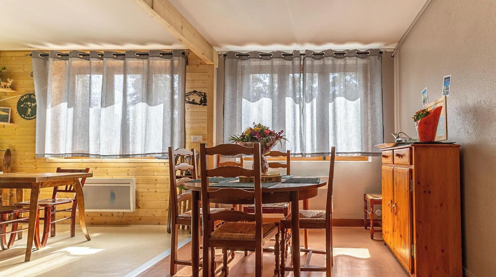 A cozy room with a wooden dining table, chairs, a cabinet, and two windows with curtains.