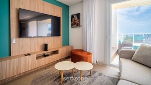 A modern living room with a flat-screen TV mounted on a wooden wall, a small orange ottoman, two round tables, and a sofa. - Penthouse With Ocean View Ingleses Mhc0306 (Florianópolis)