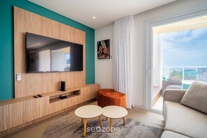 A modern living room with a flat-screen TV mounted on a wooden wall, a small orange ottoman, two round tables, and a sofa. - Penthouse With Ocean View Ingleses Mhc0306 (Florianópolis)