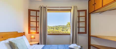 A bedroom with a bed, a nightstand, a lamp, a window with curtains, and a view of greenery outside.