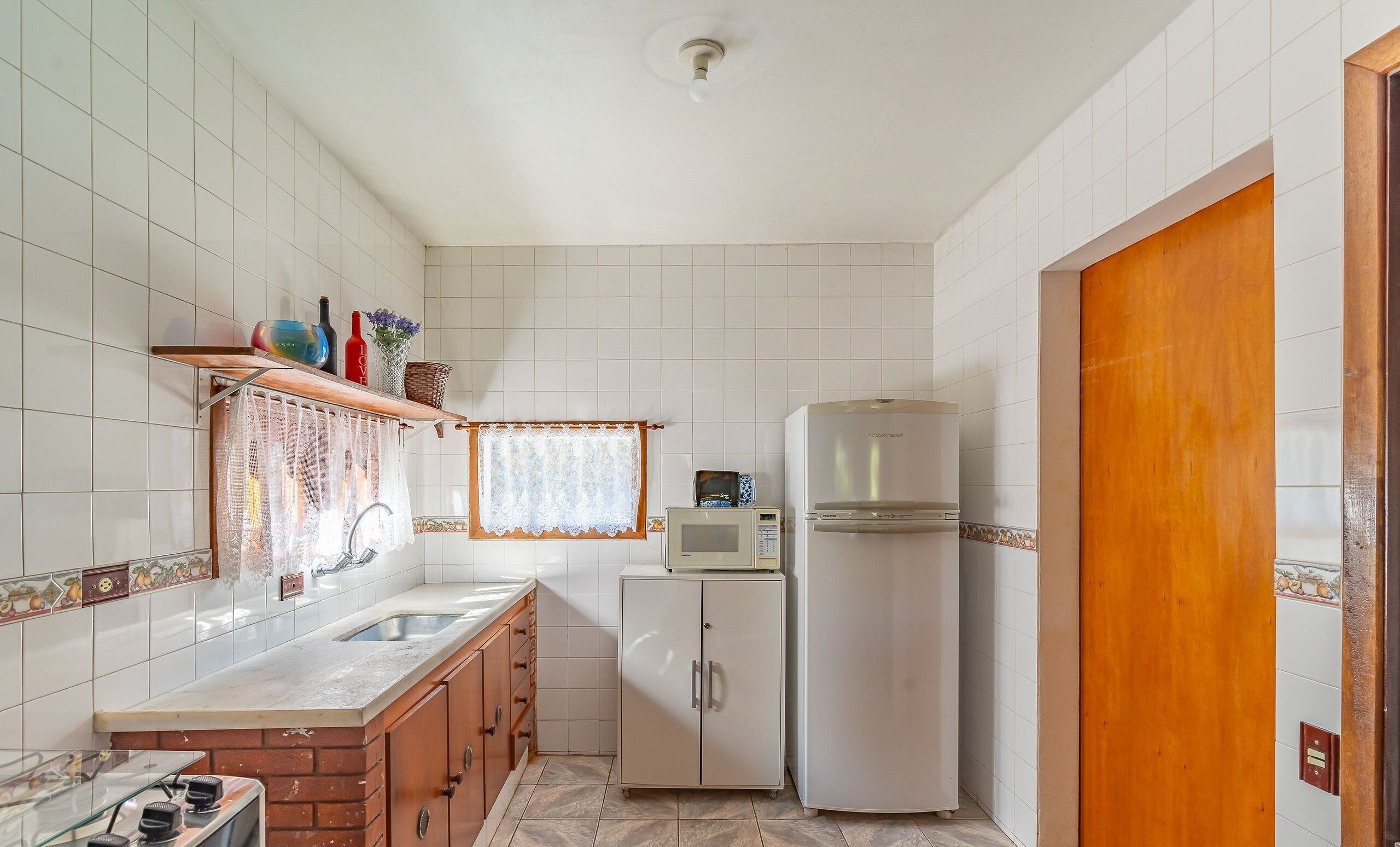 A compact kitchen with white tiles, a wooden cabinet, a microwave, and a refrigerator.